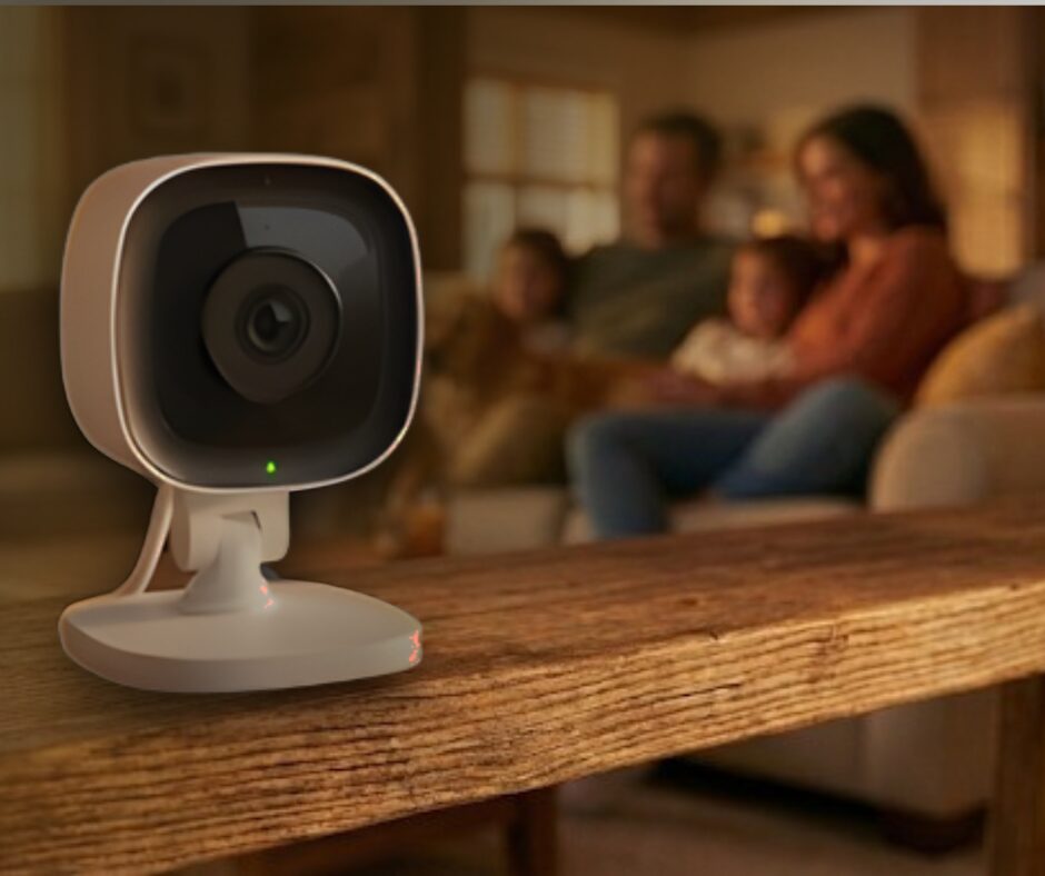 A smart indoor security camera with its mechanical privacy shutter closed, sitting on a shelf while a family relaxes in the background, illustrating home privacy.